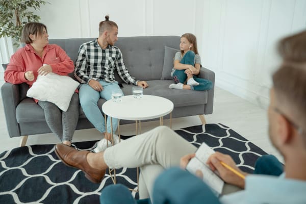 A family therapy session with a woman in a red shirt, a man in a checkered shirt, and a young girl sitting on a gray couch, while a therapist takes notes in the foreground.